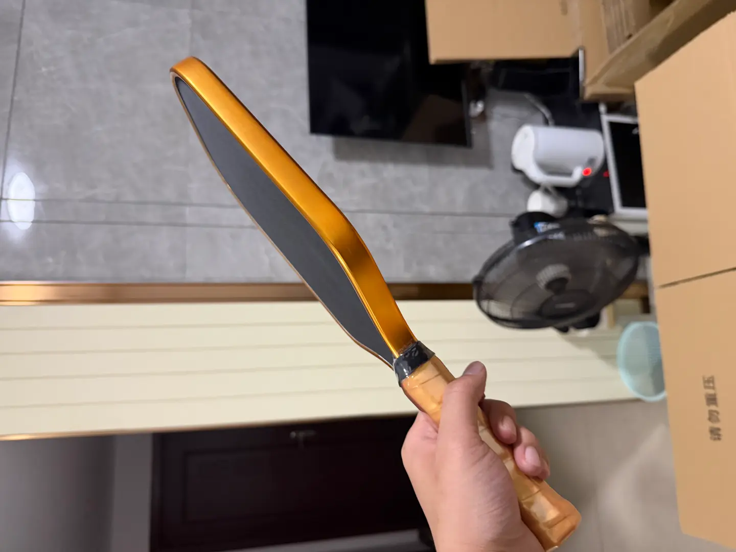 Project photograph of a dark-faced paddle held at an angle, showing its gold-colored edge and wrapped grip