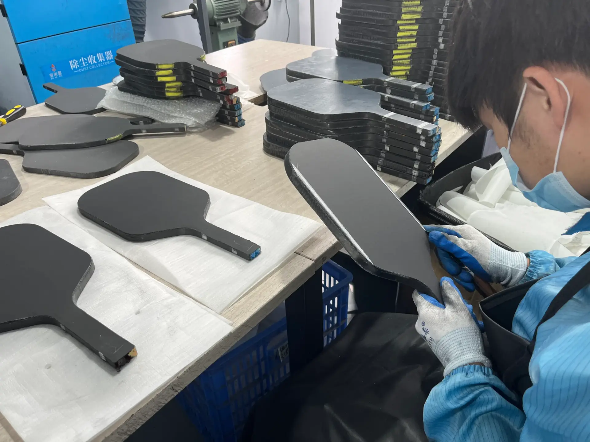 Worker handling an unfinished pickleball paddle at a workbench with stacked paddle blanks